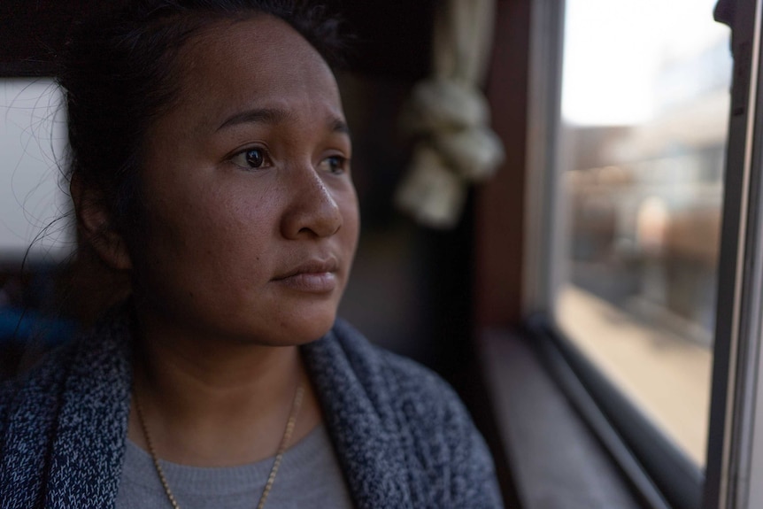 Pisey Eng looks out a window with a tearful expression. She wears a wool cardigan, has a gold necklace on and hair tied back.