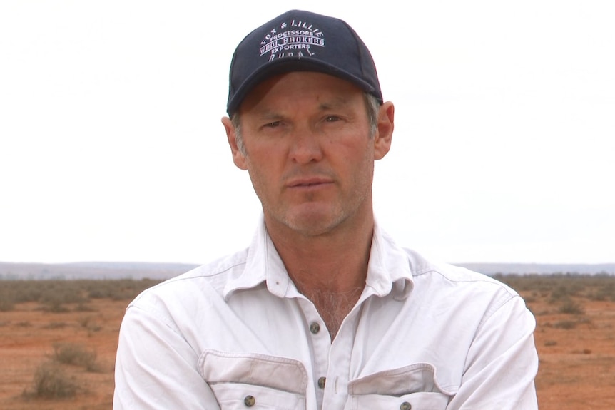 A man with a white shirt and a blue hat stands with arms folded. 