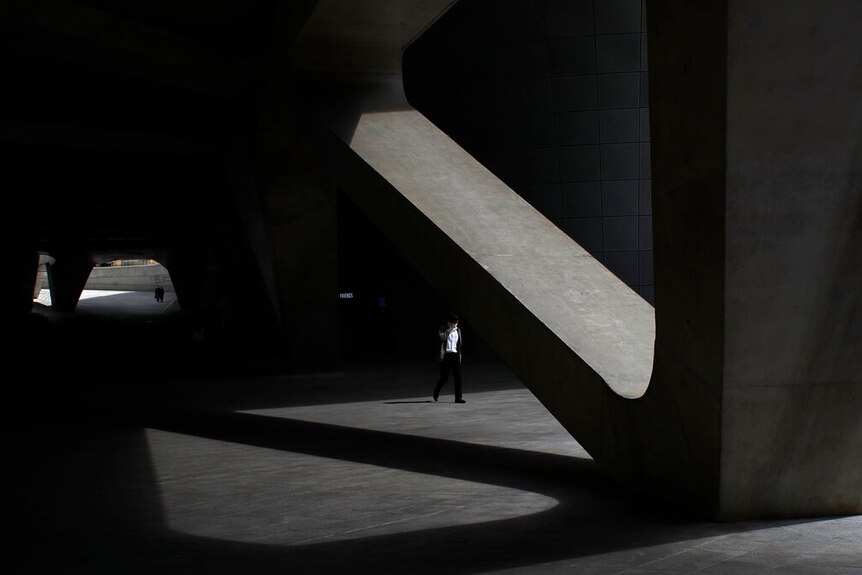 A man wearing a face mask walks amid an austere concrete square casting long shadows.