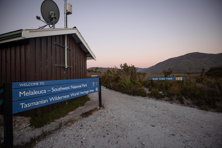 A wooden vertical board hut with a sign saying 'welcome to Melaleuca" on one side, and a sign saying 'South Coast Track nearby