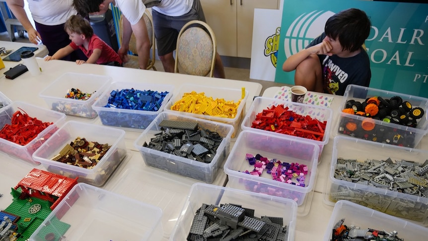 Boxes of Lego on a table surrounded by kids.