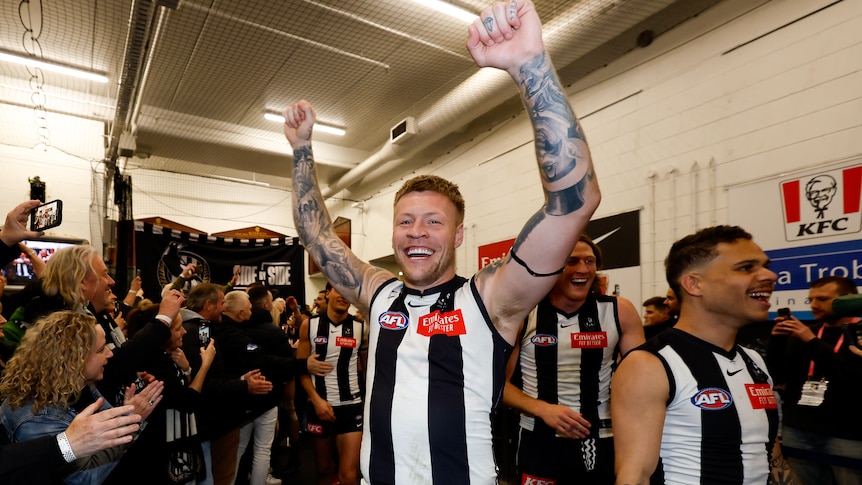 Jordan De Goey holds his arms up and smiles in the Collingwood dressing room