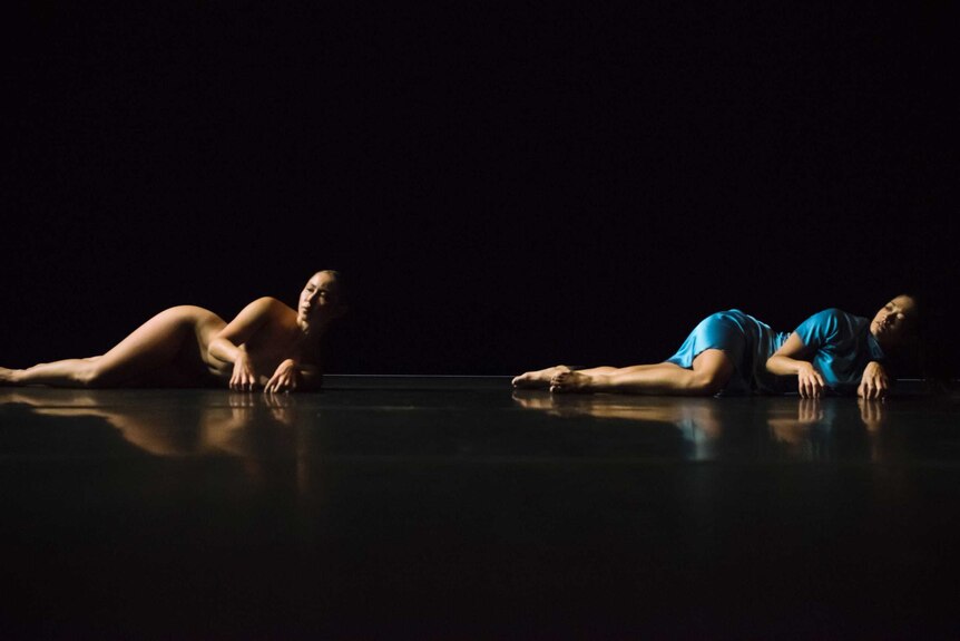 On a black stage with black background, two female dancers lie sideways - one in blue robe lies and the other is naked