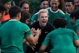 A film still of Michael Fassbender, crouching and screaming, surrounded by a Pacific Islander football team, in green.