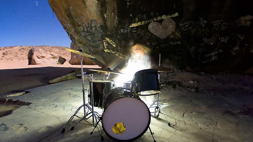 Tina is sitting behind a drumkit, blurred by movement and light. Behind her is a giant rock covered in graffiti and a desert sky