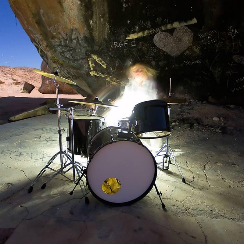 Tina is sitting behind a drumkit, blurred by movement and light. Behind her is a giant rock covered in graffiti and a desert sky
