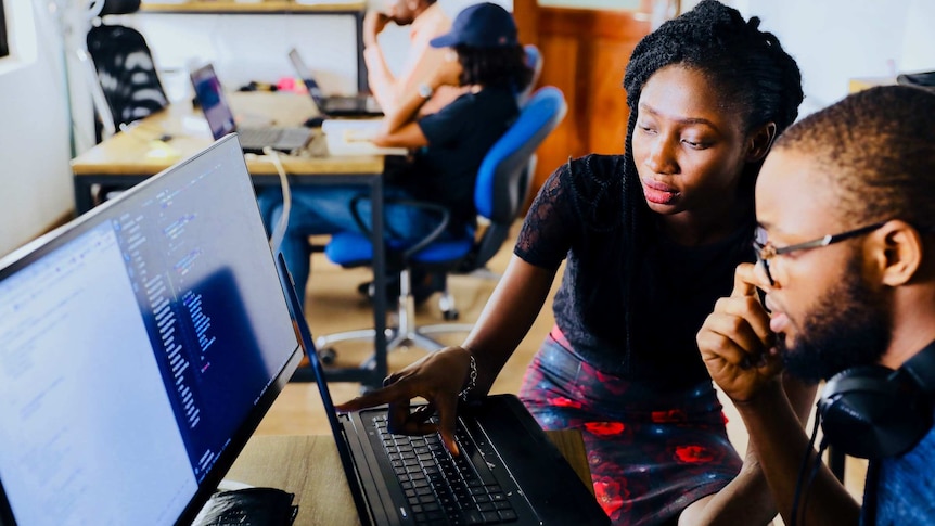 A man and a woman sit around a computer in an office, the woman is pointing to the screen