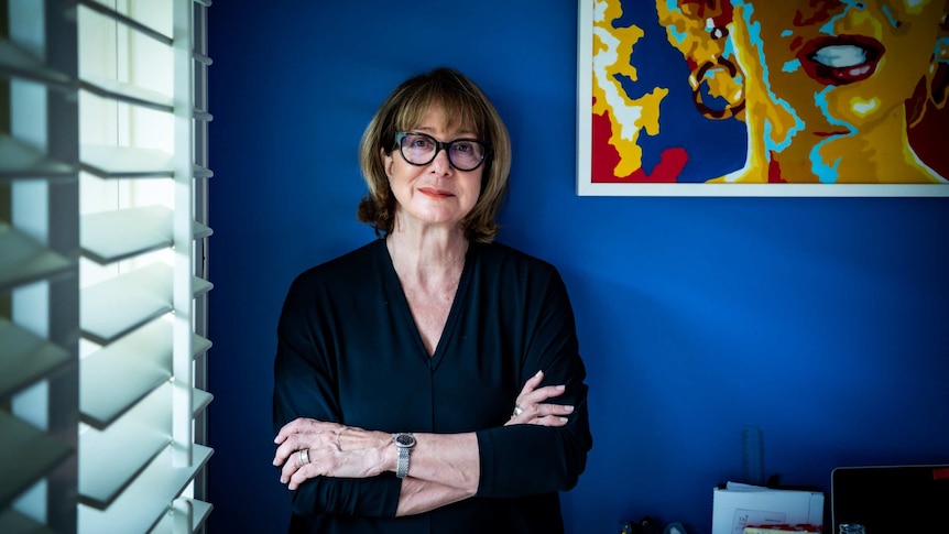 A woman with black glasses up against a blue wall