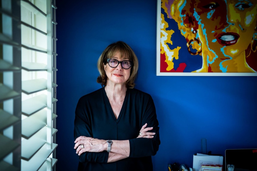 A woman with black glasses up against a blue wall
