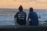 Two people sitting watching surfers, seen from behind — the woman's T-shirt says "Anti social social club"
