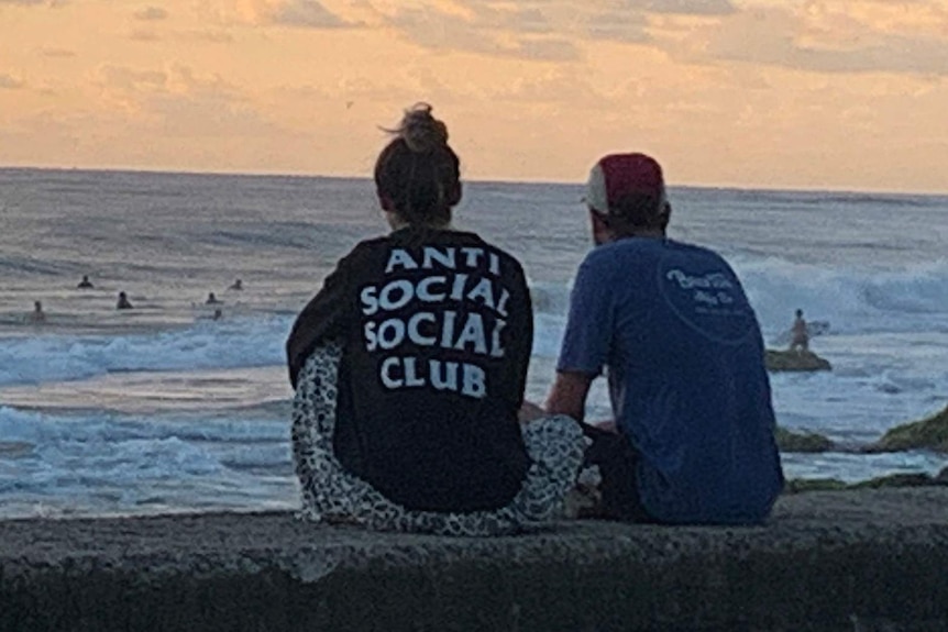 Two people sitting watching surfers, seen from behind — the woman's T-shirt says "Anti social social club"