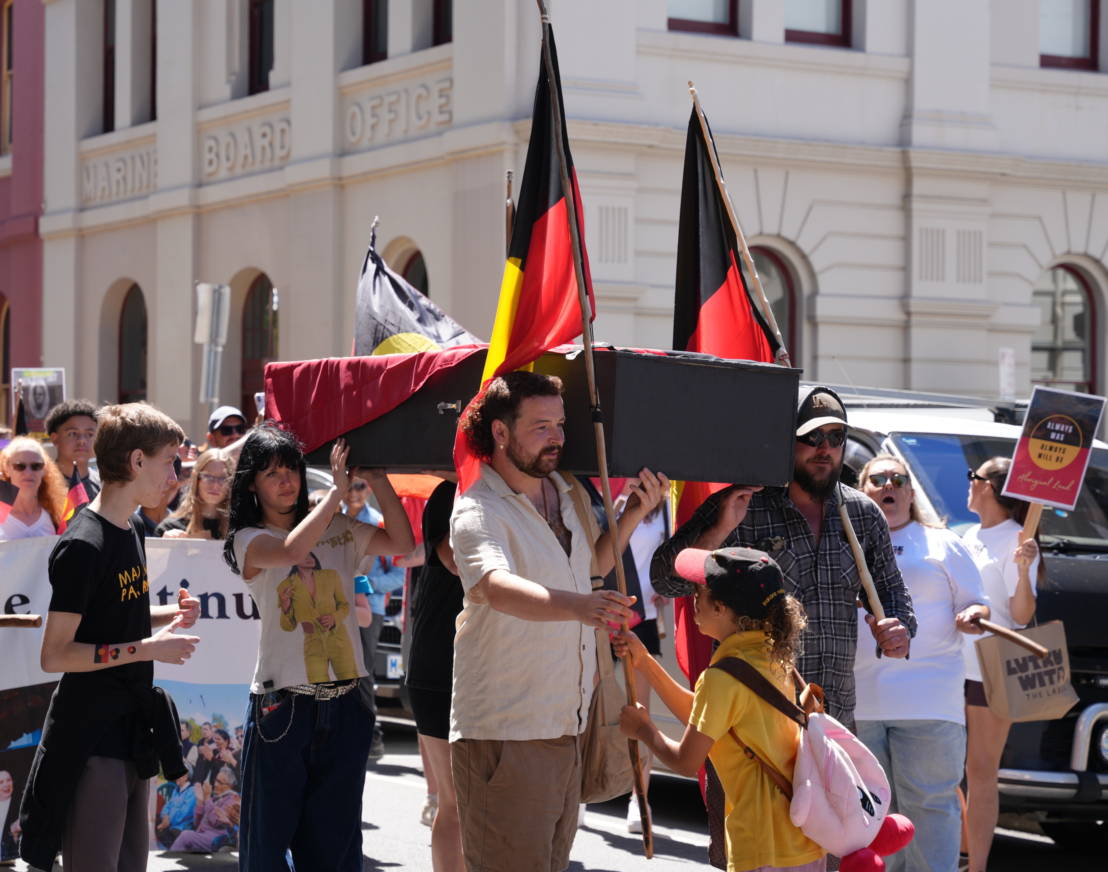 A man takes an Aboriginal flag from a young girl wearing yellow, he holds a coffin, a crowd gathers behind them