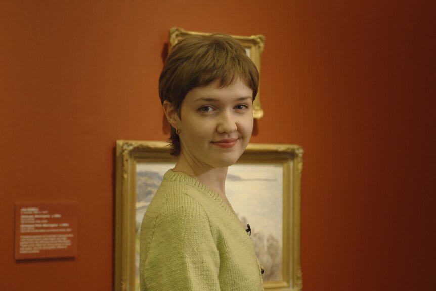 Mary McGillivray, a young woman with a short haircut, standing in front of a painting and red wall in a gallery