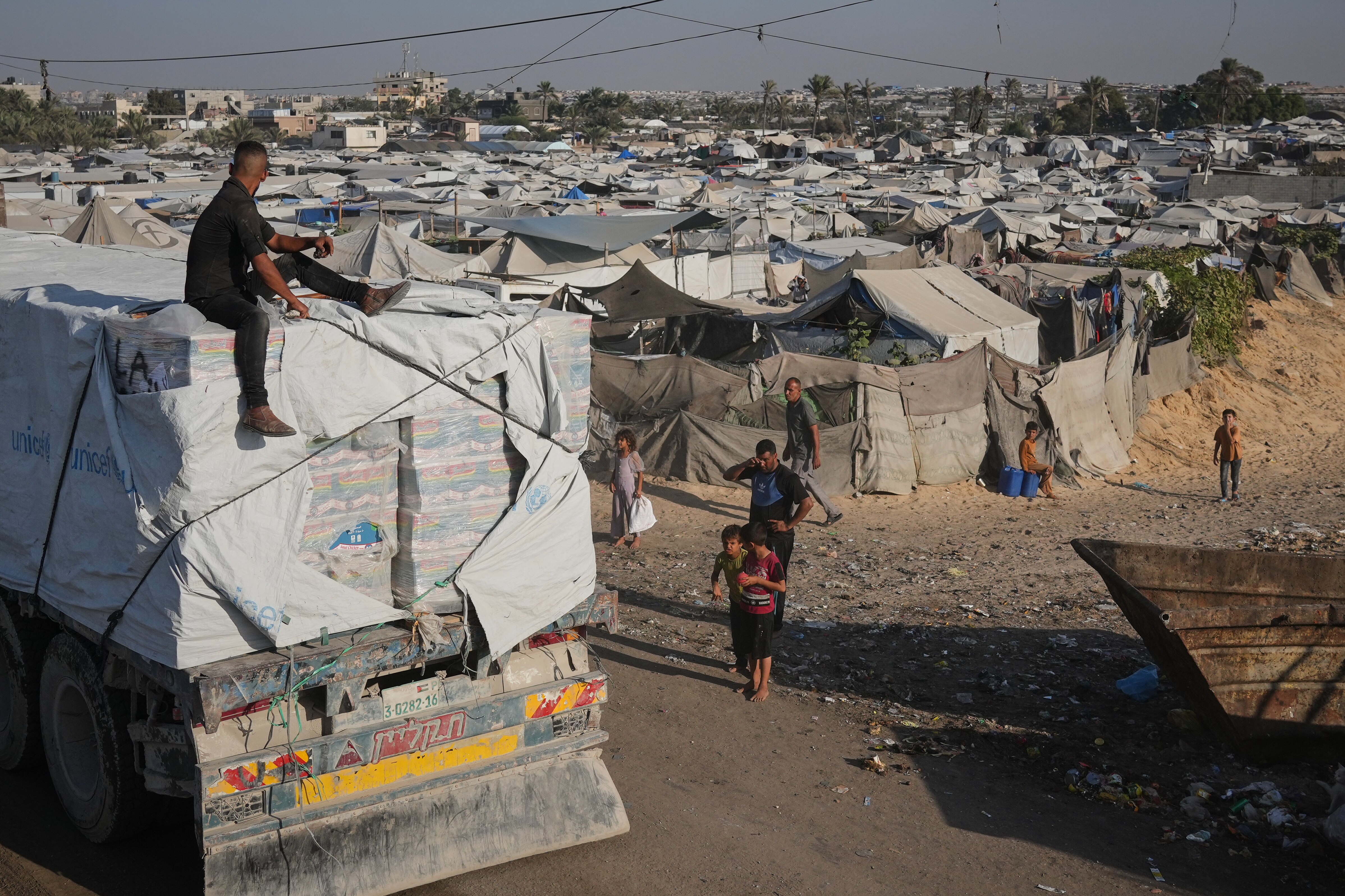 Palestinians walking next to a large tent city and a humanitarian aid truck covered in white sheets, with a man sitting on top