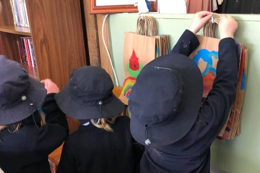 Three young school students pictured from behind with navy jumpers and hats, hanging colourful paper bags on hooks.