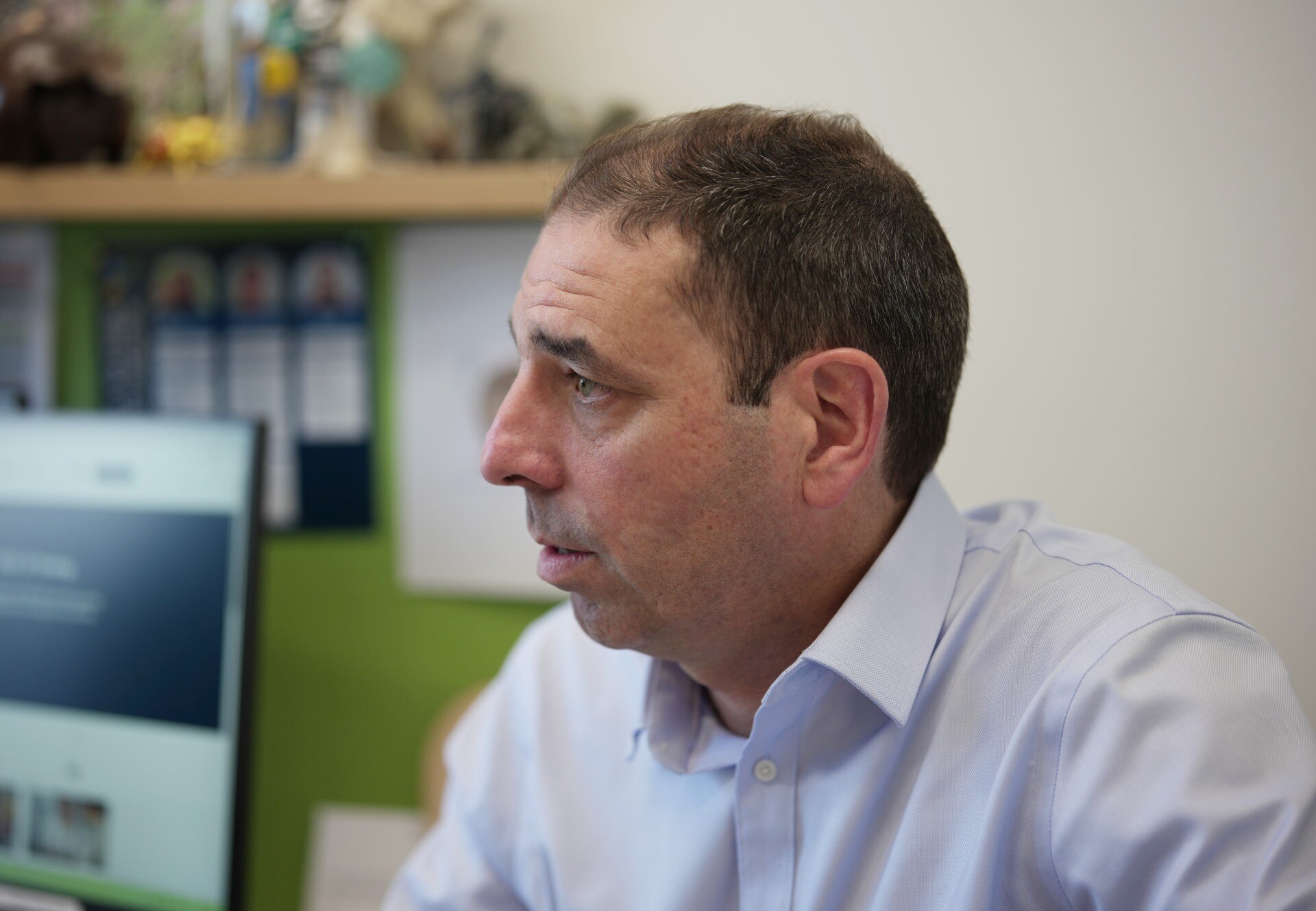 A man wearing a grey shirt sits in an office with a computer screen visible in the background. 