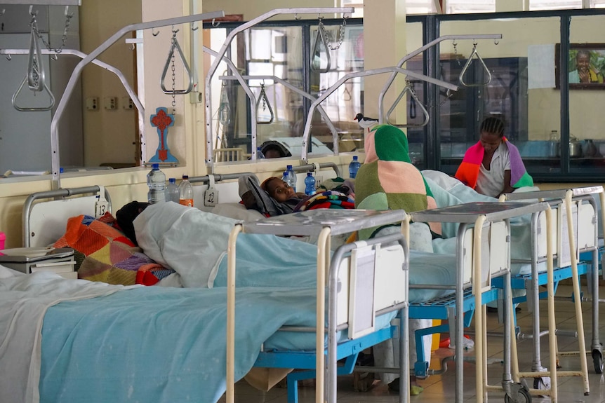 Several women lie on beds with blue blankets draped over them. Some women also have brightly-coloured knitted blankets.