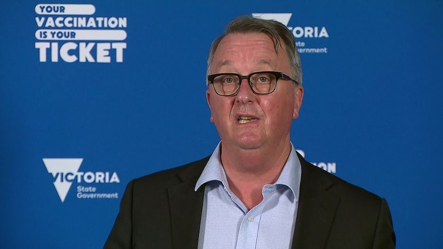 Martin Foley at a press conference, in front of a blue Vic Gov backdrop that reads 'your vaccination is your ticket'