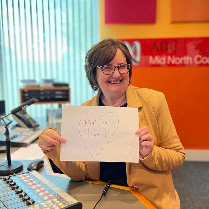 Smiling woman in radio studio holding up sign with a love heart and the question what is love.