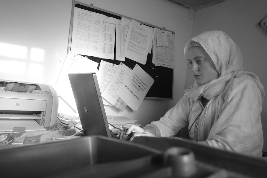 A woman in a hijab works on a laptop computer next to a cluttered pin board