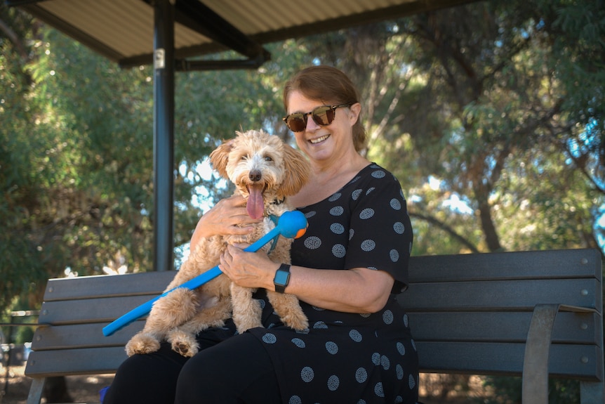 a woman sits on a park bench smiling with a dog in her lap