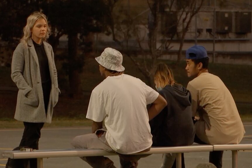 A young blonde-haired woman in black shirt and tan coat talks to three youngster sitting on bench.