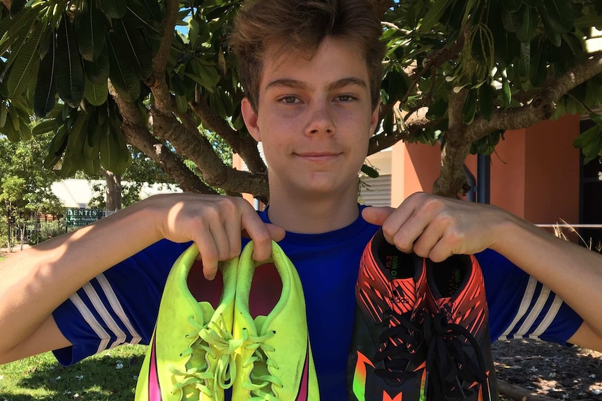 Max Beadle holding up two sets of footy boots in front of tree
