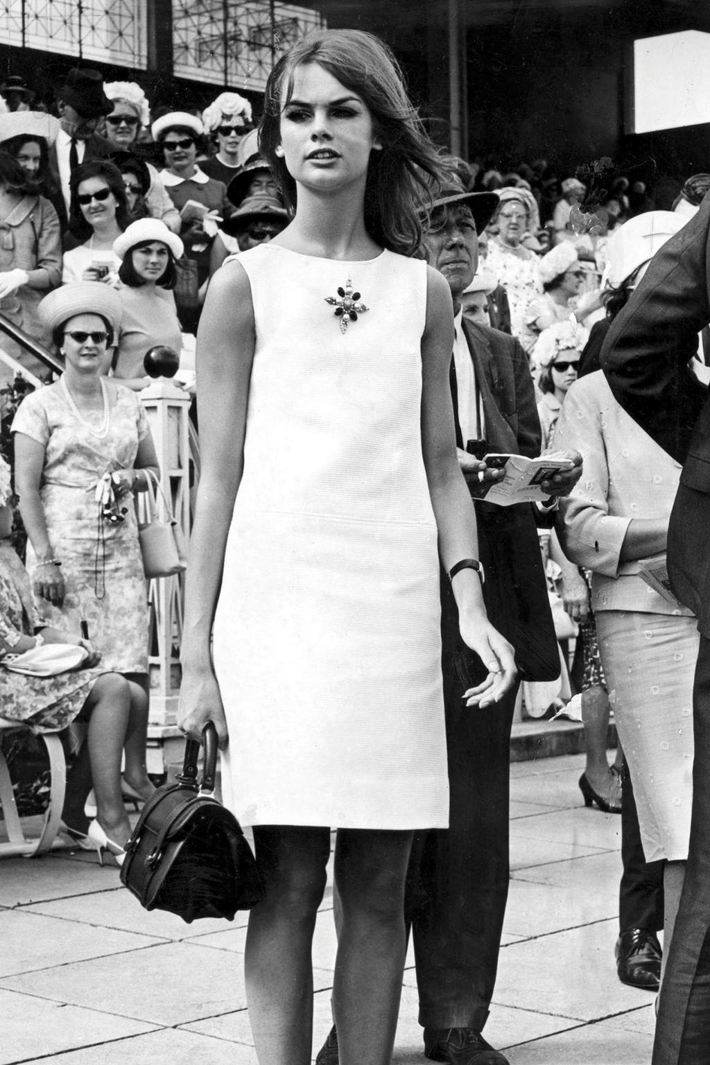 A black-and-white photograph of Jean Shrimpton in a white minidress at the races.