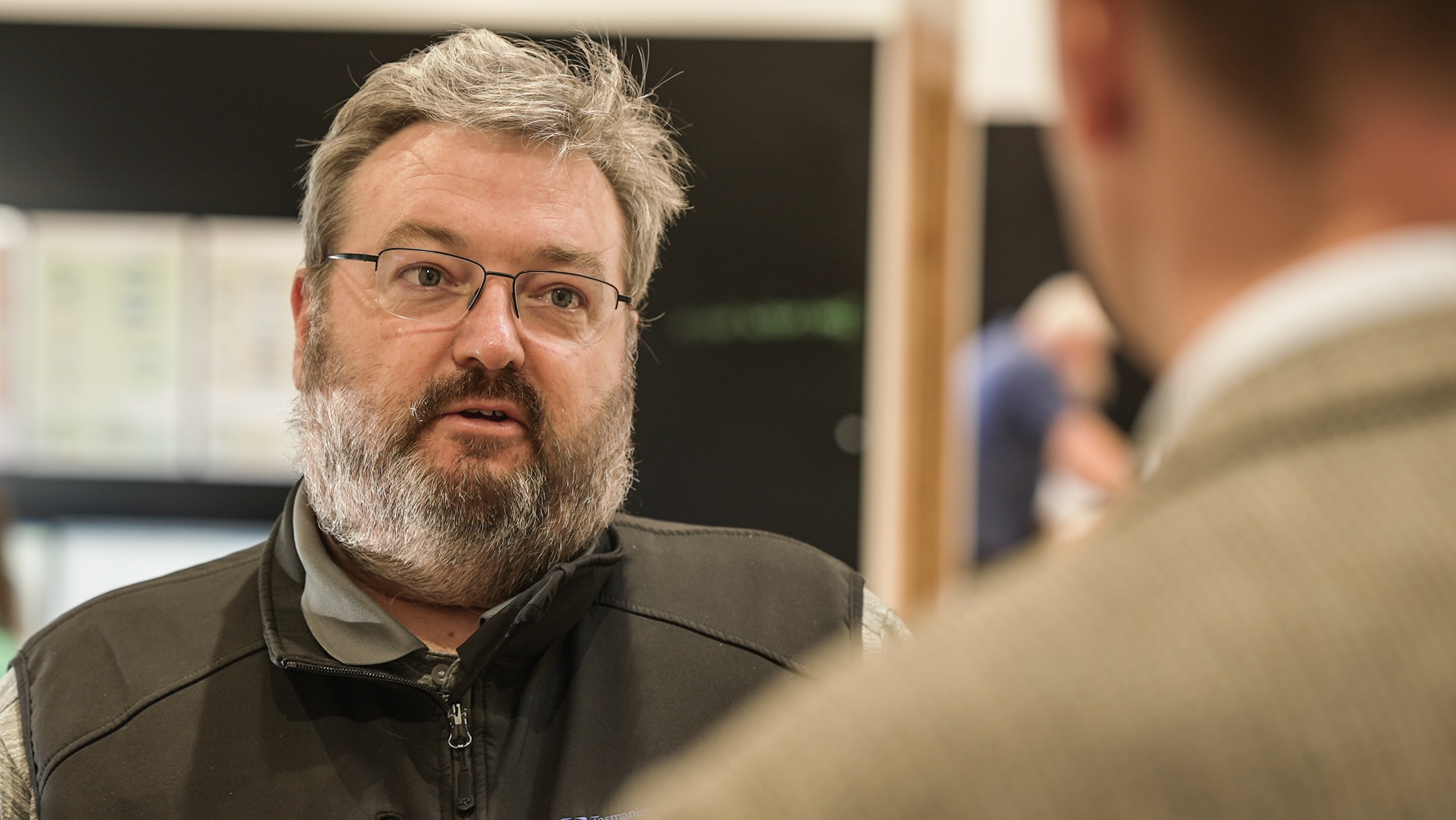 Bearded man wearing glasses speaking to out of focus person