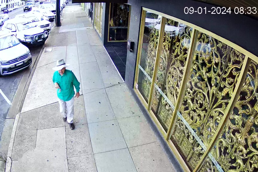 CCTV footage of a man in green shirt wearing white hat walks along the footpath of a main street