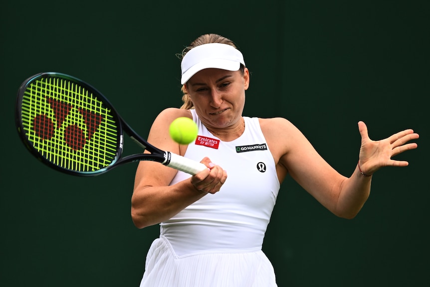 A white female players wearing a white cap and sleeveless white top plays a forehand at a ball directly in front of her.