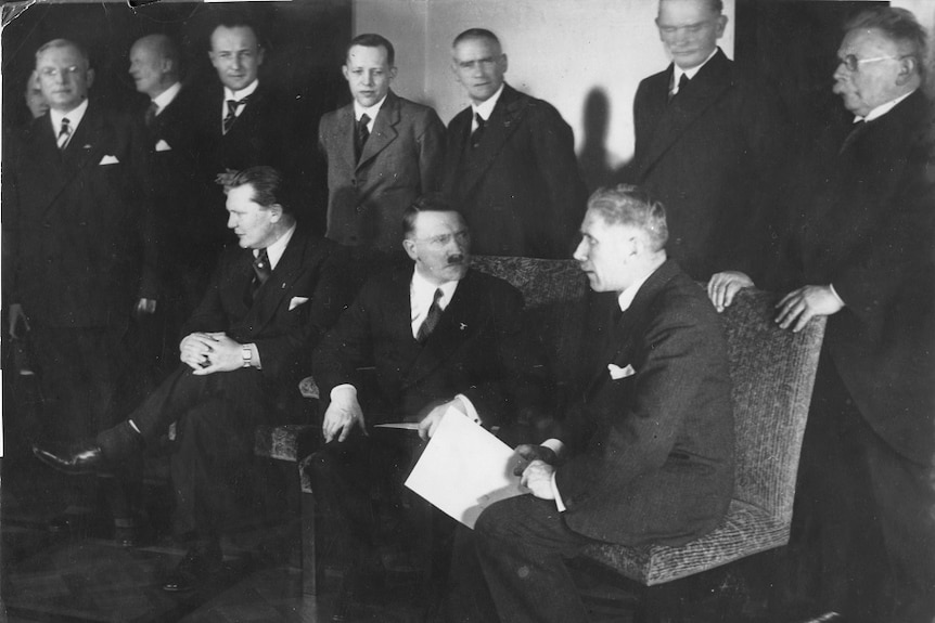 A dozen men in suits stand and sit in a line