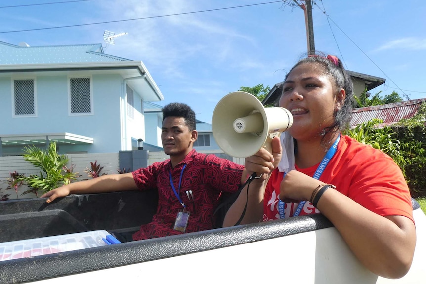 Two medical staff sit in the back of a ute, with one holding up a megaphone to her mouth.