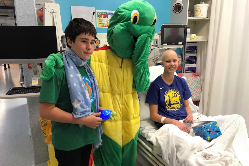 A young patient smiles in a hospital bed. A boy and a person dressed up as a giant turtle stand next to her.