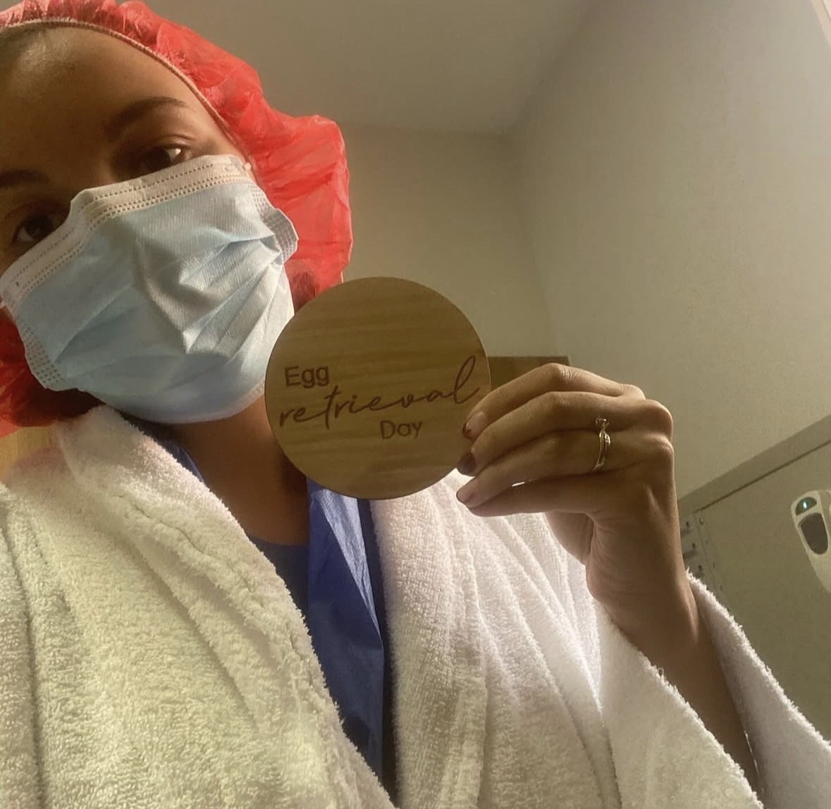 A woman wearing medical scrubs and a cap holds a small sign which reads 'egg retrieval day'.