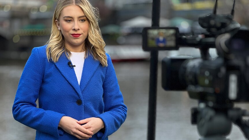 Blonde hair woman in her 20s wearing a white shirt and blue coat. She is addressing the camera.