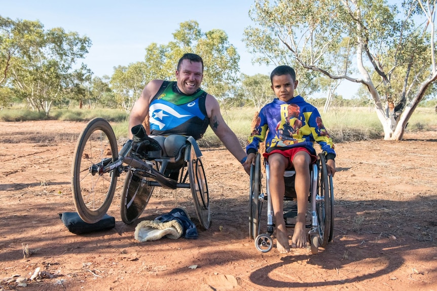 Kurt Fearnley in his racing chair smiles as he sits next to young Duqarn in Kurt's wheelchair, on red dirt.