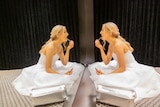 A blonde-haired girl in a white dress studies her reflection in a mirror.
