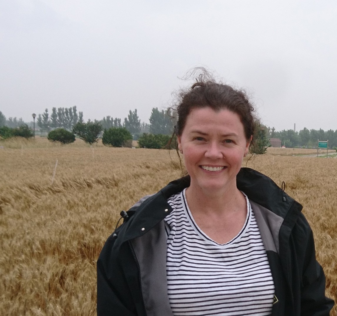 A woman, wearing a jacket, standing in a field while smiling at the camera.