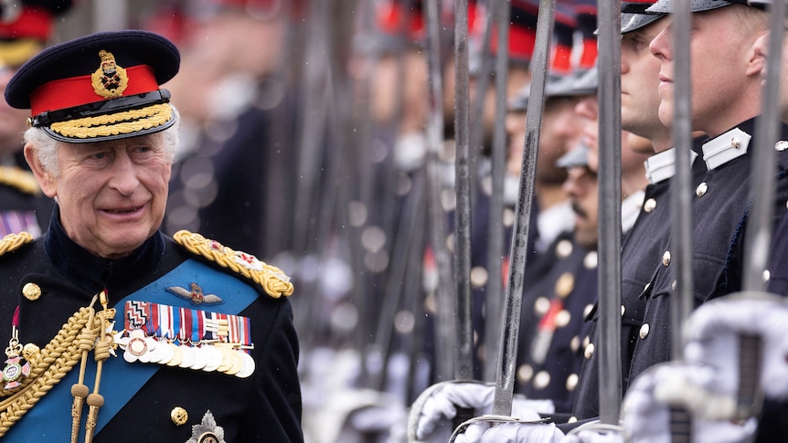 King Charles walks past a line of men holding their swords upwards. He is wearing a royal uniform. 