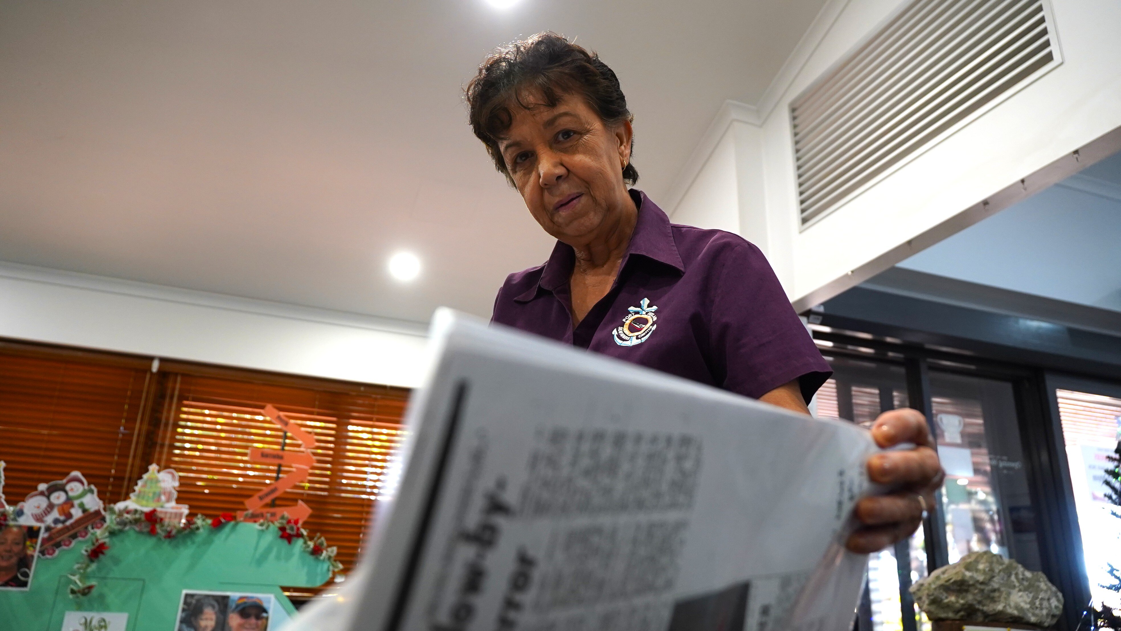 Una mujer mayor con piel bronceada hojea un viejo recorte de periódico y mira a la cámara.