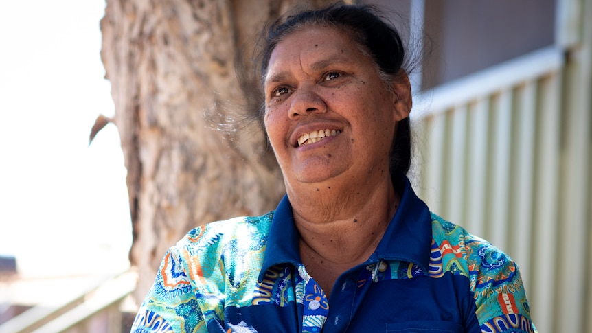 A smiling woman in a blue t-shirt featuring Aboriginal designs, standing in front of a tree. 