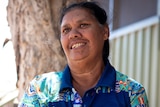 A smiling woman in a blue t-shirt featuring Aboriginal designs, standing in front of a tree. 