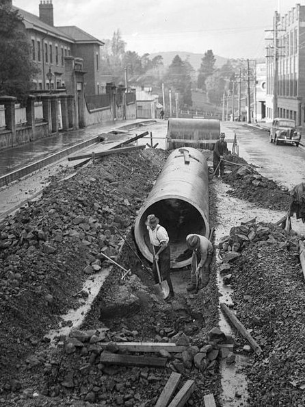 A black and white image of two men digging in a road with a large cement pipe in the background