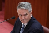 Sitting on a red seat, Mathias Cormann looks across the room.