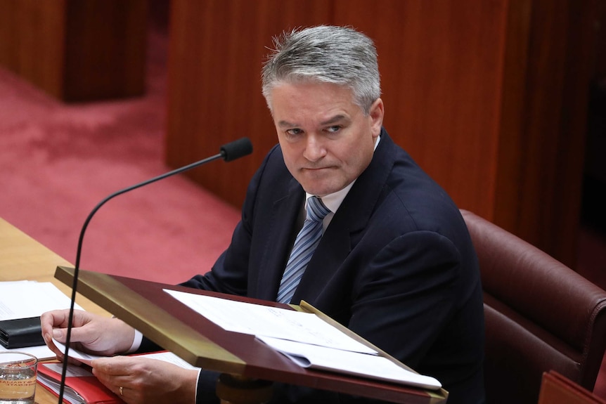 Sitting on a red seat, Mathias Cormann looks across the room.