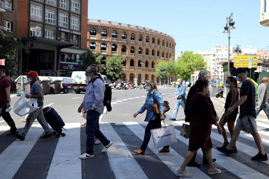 People wearing face masks walk across a pedestrian crossing in front of an amphitheatre