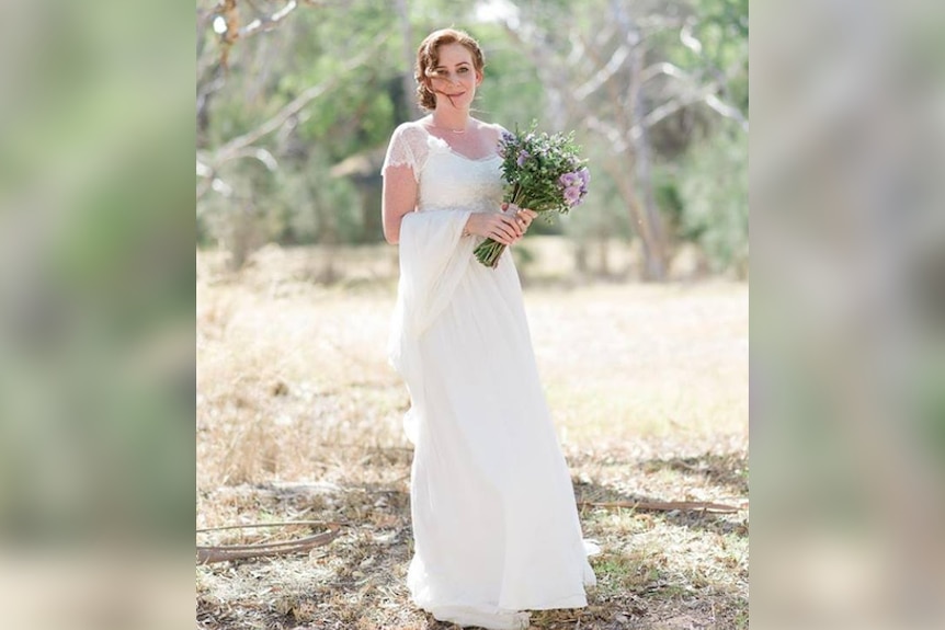 A woman with auburn hair stands outside in a wedding dress.