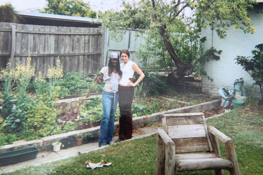 Two women are seen standing in a backyard with vegetable gardens behind them. They wear flared jeans and t-shirts.