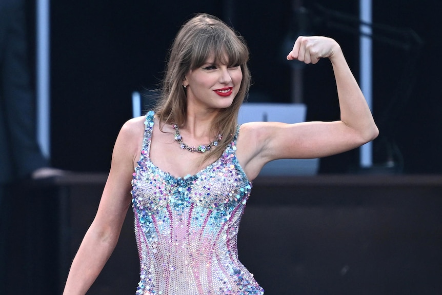 Taylor Swift flexing her arm muscle on stage. She's wearing a pastel, sequined leotard.